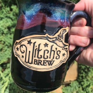 Könnte beinhalten: Schwarze Keramiktasse mit Henkel, mit einem dekorativen Etikett mit den Worten "Witch's Brew" und einem Hexenhut. Die Tasse hat eine glänzende Oberfläche mit Farbverläufen in Blau, Lila und Schwarz.