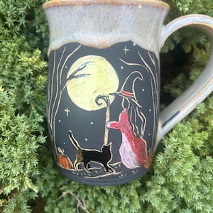 May include: A ceramic mug with a matte black surface featuring a hand-painted scene of a witch in a red cloak and hat standing with a staff beside a black cat under a large yellow moon. The mug has a light tan, speckled glazed rim and handle.