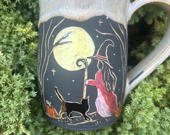 May include: A ceramic mug with a matte black surface featuring a hand-painted scene of a witch in a red cloak and hat standing with a staff beside a black cat under a large yellow moon. The mug has a light tan, speckled glazed rim and handle.
