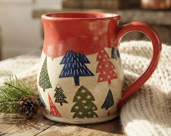 May include: A ceramic mug with a glossy red rim and handle, featuring a cream-colored body decorated with a repeating pattern of stylized pine trees in shades of blue, green, and red. The mug has a speckled texture and a rounded shape.