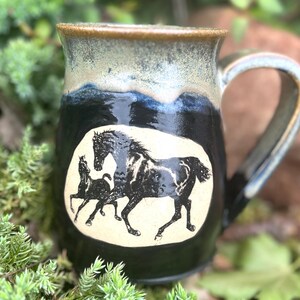 Puede incluir: Taza de cerámica hecha a mano con un diseño de caballo negro y crema. La taza tiene un borde y un asa azul oscuro y beige. La taza está sobre un lecho de follaje verde.
