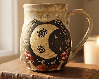 May include: A ceramic mug with a cream-colored rim and a black body featuring a gold crescent moon with spiral eyes, surrounded by white stars, leafy vines, and red-and-white spotted mushrooms at the base, with a curved handle.