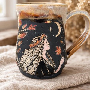 May include: A ceramic mug with a black base and a tan rim. The mug features a sgraffito illustration of a person with long hair wearing a floral crown, surrounded by falling autumn leaves, a crescent moon, and small stars against a dark background.
