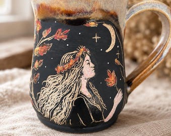 May include: A ceramic mug with a black base and a tan rim. The mug features a sgraffito illustration of a person with long hair wearing a floral crown, surrounded by falling autumn leaves, a crescent moon, and small stars against a dark background.