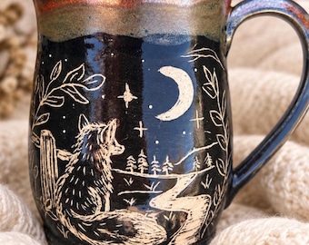 May include: A black ceramic mug with a metallic bronze rim and a handle. The surface features an etched illustration of a fox sitting near a winding path, trees, and a crescent moon with stars. The design is framed by leafy branches.
