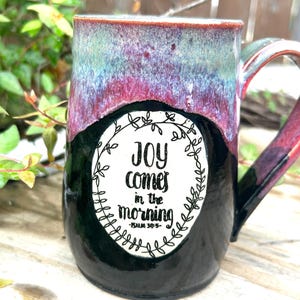 May include: A handmade ceramic mug with a black base and a colorful top. The top of the mug has a blend of blue, purple, and red hues. The mug features the text "Joy comes in the morning" inside a white floral wreath.