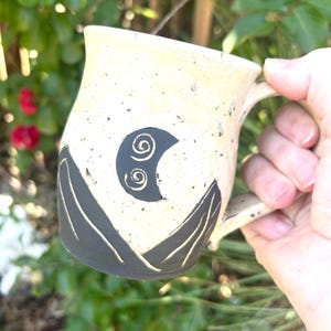 May include: A ceramic mug with a speckled cream-colored glaze featuring a dark gray mountain range design at the base and a dark gray crescent moon with two spiral patterns in the center of the body, complete with a matching speckled handle.