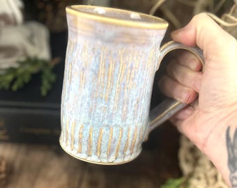 May include: A cylindrical ceramic mug featuring a textured surface with vertical ridges. The glaze is a blend of soft blue and lavender tones, with warm tan or beige accents highlighting the rim, the handle, and the recessed vertical grooves.