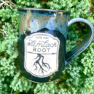 May include: A ceramic mug with a glossy finish featuring a blue and black gradient glaze. A white label on the front displays the text 100% PURE Hemlock ROOT above a black line drawing of a root system with radiating lines underneath the text.
