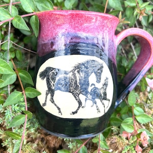 Puede incluir: Una taza de cerámica hecha a mano con un esmalte negro, morado y rojo. La taza presenta un óvalo blanco con un diseño de caballo negro y azul. El asa y el borde son rojos. La taza está rodeada de follaje verde.