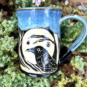 May include: A ceramic mug featuring a glossy blue glaze on the upper portion and a cream-colored base. The body displays a black sgraffito illustration of a crow perched in front of a large, circular moon, surrounded by dark, stylized tree branches.