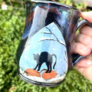 Op de afbeelding: Handgemaakte keramische mok met een zwart katten silhouet op een besneeuwde landschapsscène. De mok heeft een donkerblauwe en zwarte glazuur met een handvat. De kat staat op twee oranje pompoenen.