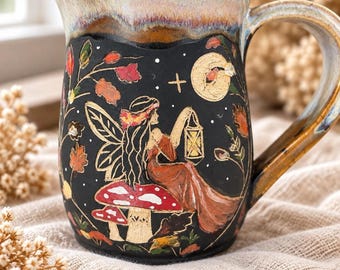May include: A ceramic mug with a black exterior featuring a hand-painted scene of a winged figure sitting on a red and white mushroom, holding a lantern under a moon and stars. The mug has a wide handle and a mottled tan and brown rim.