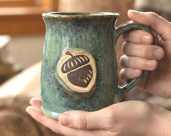 May include: A ceramic mug with a mottled green and blue glaze featuring a raised, brown, carved acorn emblem on the front. The mug has a sturdy handle and a wide rim, showcasing a textured, handcrafted appearance with visible colour variations.