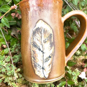 May include: A brown ceramic mug with a large handle. The mug features a painted feather design in shades of brown, tan, and black. The mug is sitting on a bed of green foliage.