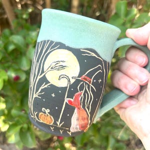 Puede incluir: Una taza de cerámica con exterior negro mate que presenta una escena pintada a mano de una luna llena amarilla, árboles desnudos, una calabaza y una figura con sombrero puntiagudo. El borde superior y el asa tienen un esmalte verde menta claro.