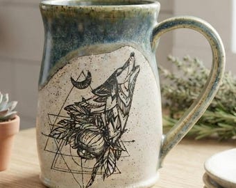 May include: A ceramic mug with a speckled cream-colored body and a blue-green glazed rim. The front features a black line-art illustration of a wolf howling at a crescent moon, integrated with geometric shapes and floral elements.