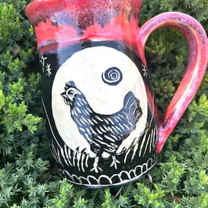 May include: A black and white ceramic mug with a red glaze. The mug features a black and white illustration of a rooster in front of a full moon. The moon has a spiral design on it.