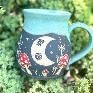 May include: A ceramic mug with a teal rim and handle, featuring a dark blue body decorated with a white crescent moon, red and white mushrooms, and botanical details. The mug is set against green foliage.