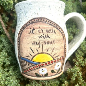 May include: Cream-colored ceramic mug with a handle. The mug features a hand-painted design with the words "it is well with my soul" above a sunrise scene. The design includes a yellow sun, a blue river, and white flowers.