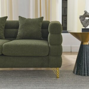 May include: Olive green boucle sofa with matching throw pillows. The sofa features a rounded armrest design and gold-colored legs. The sofa sits on a gray and white striped rug. A side table with a gold top and marble base is next to the sofa.