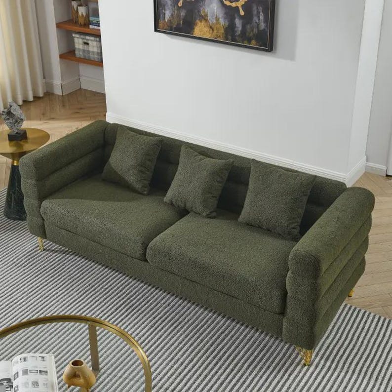 May include: A green sofa with three matching throw pillows. The sofa has a textured fabric and gold-colored legs. The sofa is placed on a striped rug.