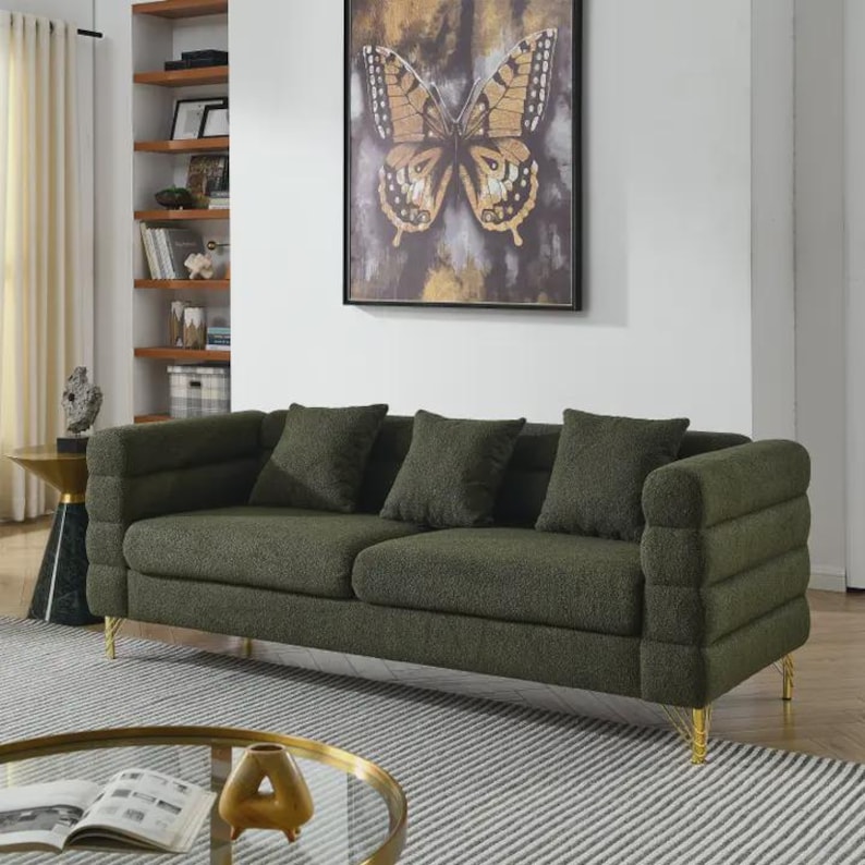 May include: A green sofa with three matching throw pillows. The sofa has gold-colored legs and a textured fabric. A framed butterfly art piece hangs on the wall behind the sofa. The sofa is on a striped rug.
