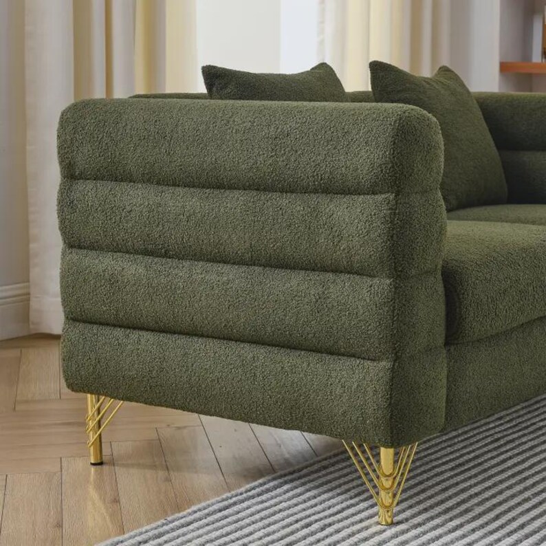 May include: Olive green sofa with a textured, ribbed design on the armrest. The sofa has gold-colored metal legs. Two matching throw pillows are visible. The sofa is placed on a striped rug.