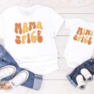Può includere: Una maglietta bianca con il testo "Mama Spice" in lettere arancioni e gialle. Un body bianco con il testo "Mini Spice" in lettere arancioni e gialle. Entrambi i capi sono su una superficie di legno con jeans blu e scarpe da ginnastica bianche.