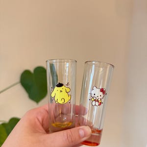 Może przedstawiać: Dwa przezroczyste kieliszki do shotów z postaciami z kreskówek. Jeden przedstawia żółtą postać przypominającą psa z czarnym kapeluszem, drugi białego kota z czerwoną kokardką. Spody szklanek mają pomarańczowe i żółte akcenty.