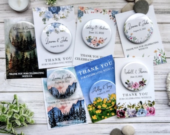 May include: Six round wedding favor magnets with watercolor designs. The magnets feature different floral and nature scenes with the couple's names and wedding date. The text on the magnets reads "Thank you for celebrating with us".