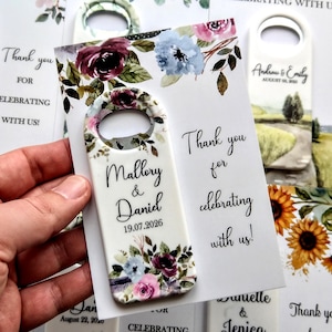 K&ouml;nnte beinhalten: Flaschen&ouml;ffner-Gastgeschenke mit floralen Designs und den Namen "Mallory & Daniel" sowie dem Datum "19.07.2026". Der Text "Thank you for celebrating with us!" ist ebenfalls sichtbar. Weitere Gastgeschenke sind im Hintergrund.