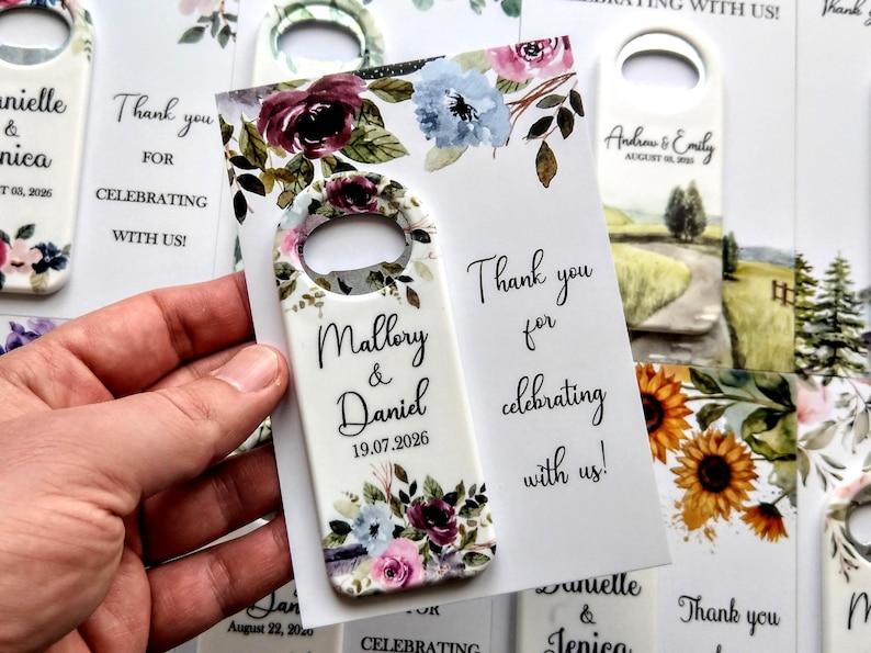 K&ouml;nnte beinhalten: Flaschen&ouml;ffner-Gastgeschenke mit floralen Designs und den Namen "Mallory & Daniel" sowie dem Datum "19.07.2026". Der Text "Thank you for celebrating with us!" ist ebenfalls sichtbar. Weitere Gastgeschenke sind im Hintergrund.