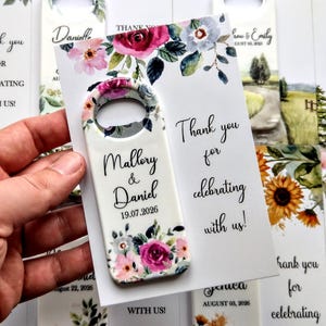 K&ouml;nnte beinhalten: Ein wei&szlig;er Flaschen&ouml;ffner mit floralem Design und den Namen "Mallory & Daniel" sowie dem Datum "19.07.2026". Der &Ouml;ffner wird neben einer Karte mit der Aufschrift "Danke, dass Sie mit uns feiern!" gehalten.