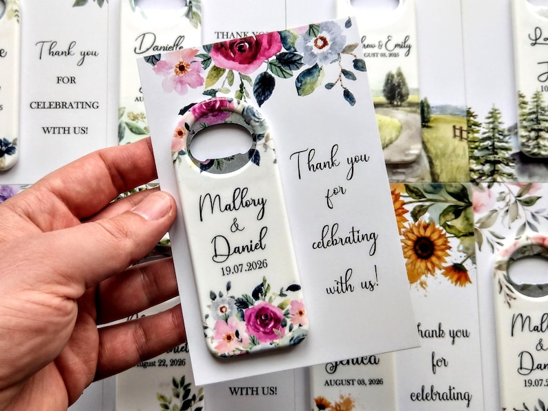 K&ouml;nnte beinhalten: Ein wei&szlig;er Flaschen&ouml;ffner mit floralem Design und den Namen "Mallory & Daniel" sowie dem Datum "19.07.2026". Der &Ouml;ffner wird neben einer Karte mit der Aufschrift "Danke, dass Sie mit uns feiern!" gehalten.