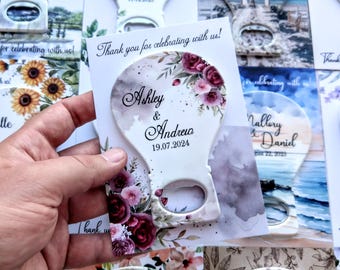 May include: A white bottle opener with a floral design and the names "Ashley & Andrew" and the date "19.07.2024". The opener is held in a hand, with other similar items in the background.