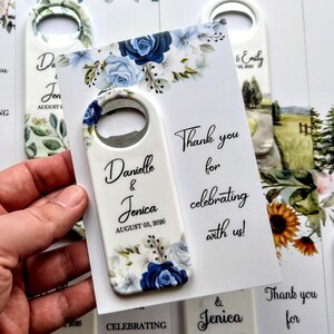 K&ouml;nnte beinhalten: Ein wei&szlig;er Flaschen&ouml;ffner mit einem blau-wei&szlig;en Blumendesign, gehalten in einer Hand. Der &Ouml;ffner tr&auml;gt die Namen "Danielle & Jenica" und das Datum "AUGUST 03, 2026". Eine Karte mit dem Text "Thank you for celebrating with us!" ist ebenfalls sichtbar.