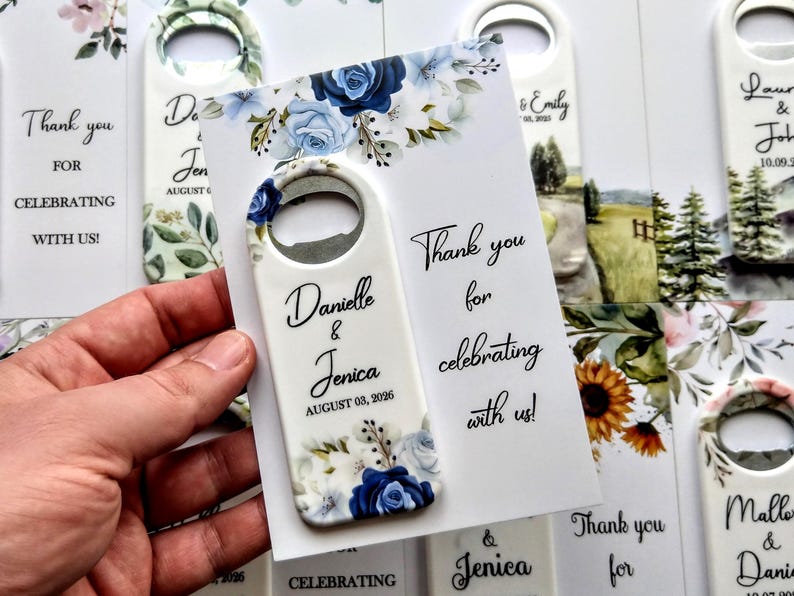 K&ouml;nnte beinhalten: Ein wei&szlig;er Flaschen&ouml;ffner mit einem blau-wei&szlig;en Blumendesign, gehalten in einer Hand. Der &Ouml;ffner tr&auml;gt die Namen "Danielle & Jenica" und das Datum "AUGUST 03, 2026". Eine Karte mit dem Text "Thank you for celebrating with us!" ist ebenfalls sichtbar.