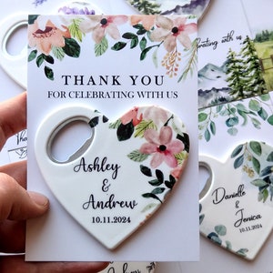 May include: A set of heart-shaped bottle openers with floral designs and personalized names and dates. The openers are white with a silver bottle opener on the side. The text on the openers reads "Ashley & Andrew 10.11.2024" and "Danielle & Jenica 10.11.2024".