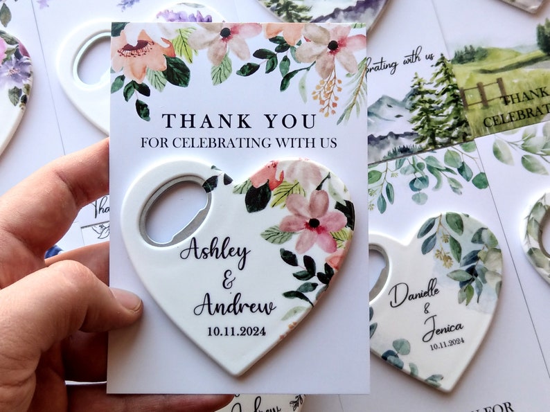 May include: A set of heart-shaped bottle openers with floral designs and personalized names and dates. The openers are white with a silver bottle opener on the side. The text on the openers reads "Ashley & Andrew 10.11.2024" and "Danielle & Jenica 10.11.2024".