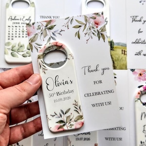 May include: White bottle opener with floral design and the text "Olivia's 50th Birthday 10.09.2026". The opener is held with a card that says "Thank you for celebrating with us!" Other openers and cards are visible.