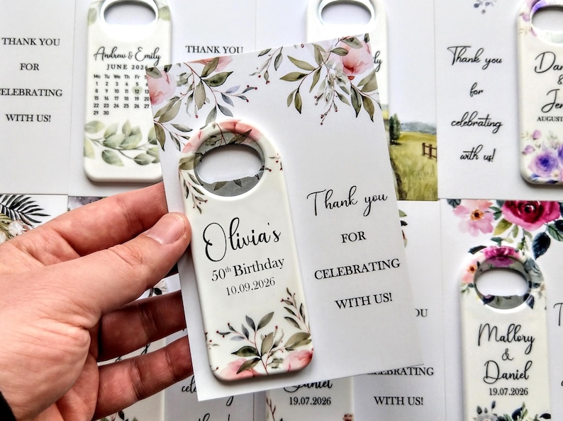 May include: White bottle opener with floral design and the text "Olivia's 50th Birthday 10.09.2026". The opener is held with a card that says "Thank you for celebrating with us!" Other openers and cards are visible.