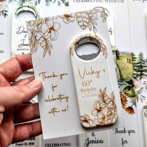 May include: A white bottle opener with a floral design and the text "Vicky's 60th Birthday 10.09.2026". The opener is held with a card that says "Thank you for celebrating with us!"