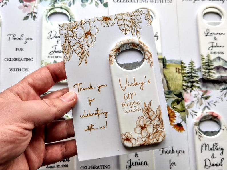 May include: A white bottle opener with a floral design and the text "Vicky's 60th Birthday 10.09.2026". The opener is held with a card that says "Thank you for celebrating with us!"