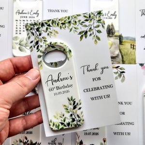 May include: A white bottle opener with a floral design and the text "Andrew's 60th Birthday 10.09.2026". The opener is held next to a card that says "Thank you for celebrating with us!"