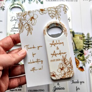 K&ouml;nnte beinhalten: Ein wei&szlig;er Flaschen&ouml;ffner mit floralem Design und den Namen "Ashley & Andrew" sowie dem Datum "19.07.2026". Daneben liegt eine Karte mit der Aufschrift "Thank you for celebrating with us!"
