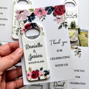 K&ouml;nnte beinhalten: Ein wei&szlig;er Flaschen&ouml;ffner mit floralem Design und den Namen "Danielle & Jenica" sowie dem Datum "03. August 2026". Der &Ouml;ffner wird mit einer Karte gehalten, auf der steht "Danke, dass Sie mit uns feiern!"