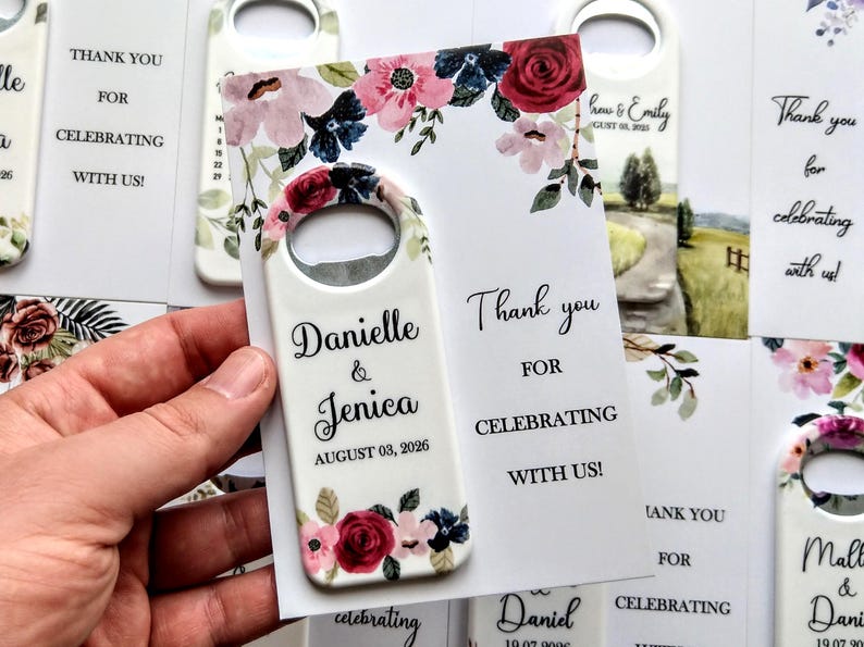 K&ouml;nnte beinhalten: Ein wei&szlig;er Flaschen&ouml;ffner mit floralem Design und den Namen "Danielle & Jenica" sowie dem Datum "03. August 2026". Der &Ouml;ffner wird mit einer Karte gehalten, auf der steht "Danke, dass Sie mit uns feiern!"