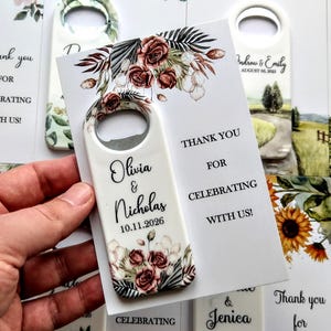 K&ouml;nnte beinhalten: Wei&szlig;er Flaschen&ouml;ffner mit floralem Design und den Namen "Olivia & Nicholas" sowie dem Datum "10.11.2026". Eine Karte lautet: "Danke, dass Sie mit uns feiern!" Hochzeitsgeschenke.