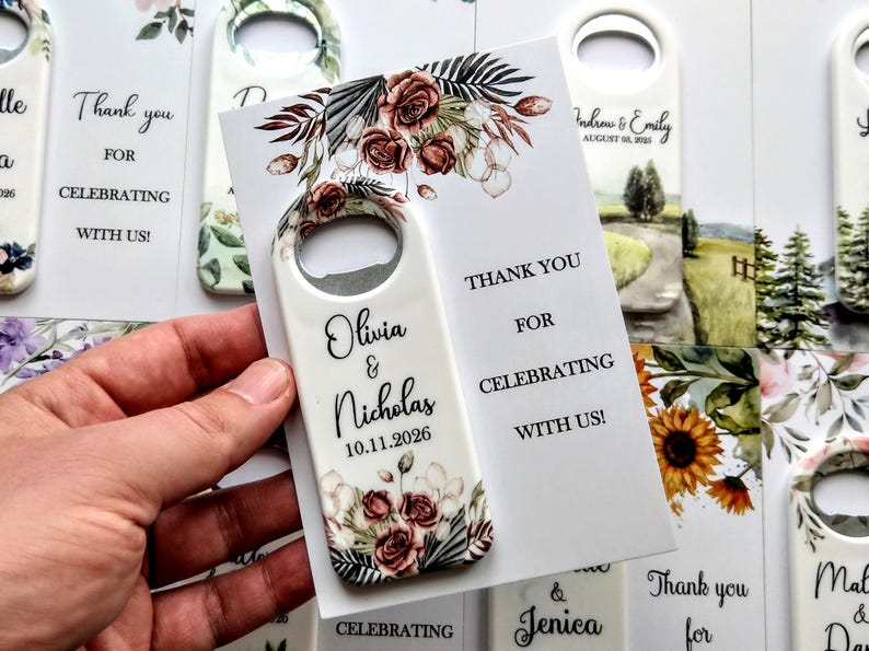 K&ouml;nnte beinhalten: Wei&szlig;er Flaschen&ouml;ffner mit floralem Design und den Namen "Olivia & Nicholas" sowie dem Datum "10.11.2026". Eine Karte lautet: "Danke, dass Sie mit uns feiern!" Hochzeitsgeschenke.
